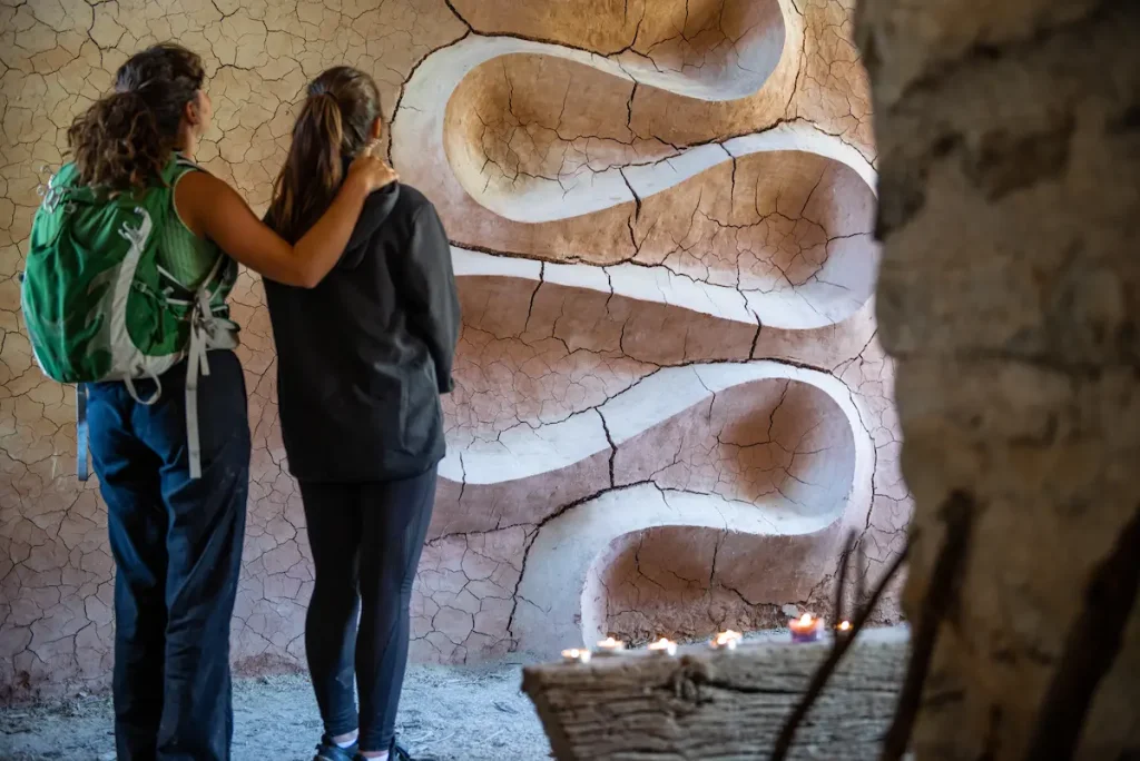 Two walkers standing before Andy Goldsworthy's cracked clay wall inside Refuge d'Art Vieil Esclangon, candles burning on a wooden beam
