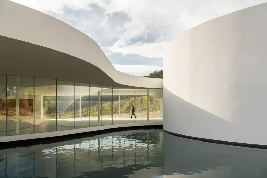 Oscar Niemeyer's white curved pavilion at Chateau La Coste, its glass wall reflected in the water basin, a visitor walking past the vineyard beyond