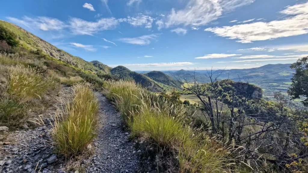 Narrow stony path traversing a grassy mountainside on the Refuge d'Art trail, the valley and the Prealps of Haute-Provence opening beyond