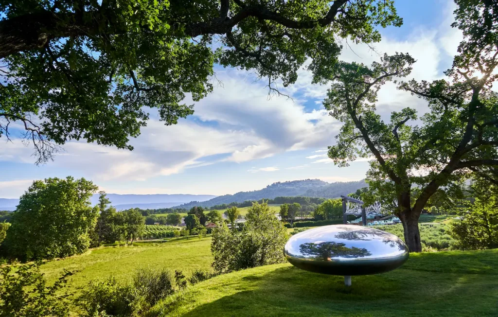Tom Shannon's mirrored sculpture Drop on the grass at Chateau La Coste, oaks framing the vineyards and a hilltop village in the evening light